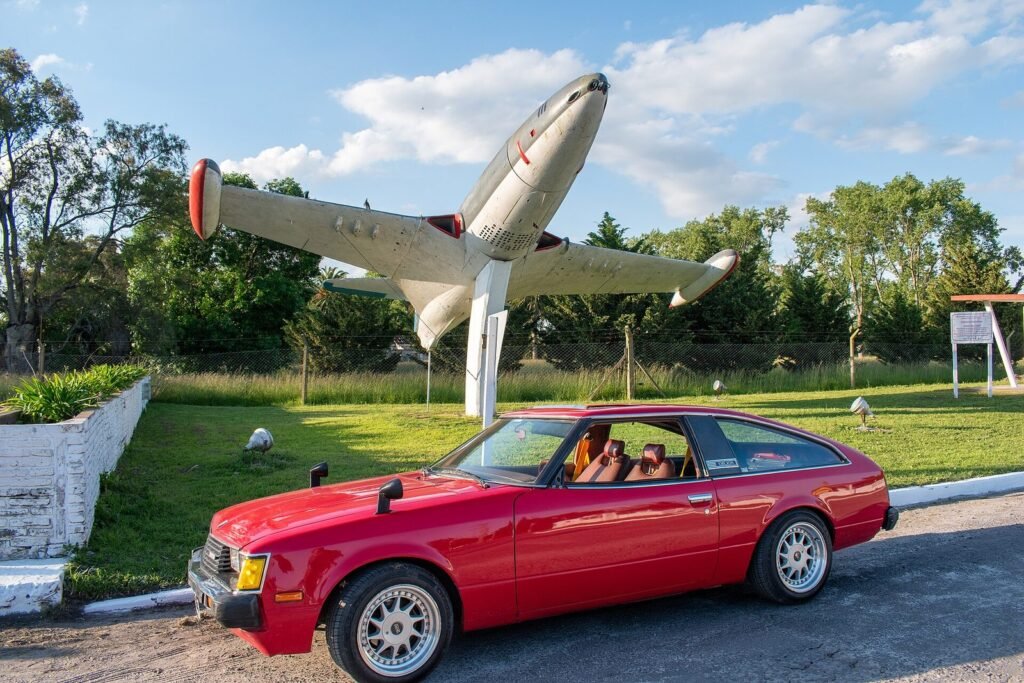 Red second-generation Toyota Celica liftback parked below a jet aircraft displayed on a pylon
