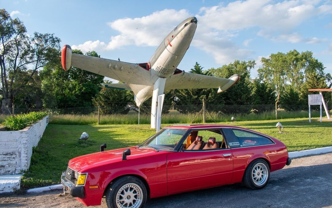 Red second-generation Toyota Celica liftback parked below a jet aircraft displayed on a pylon